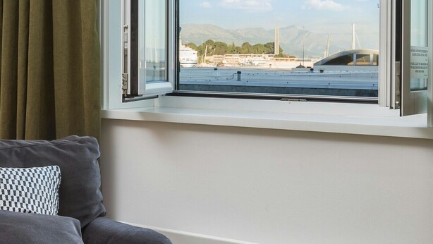 Habitació amb llit, finestra amb cortines i vista d'una marina amb vaixells i muntanyes a la distància.