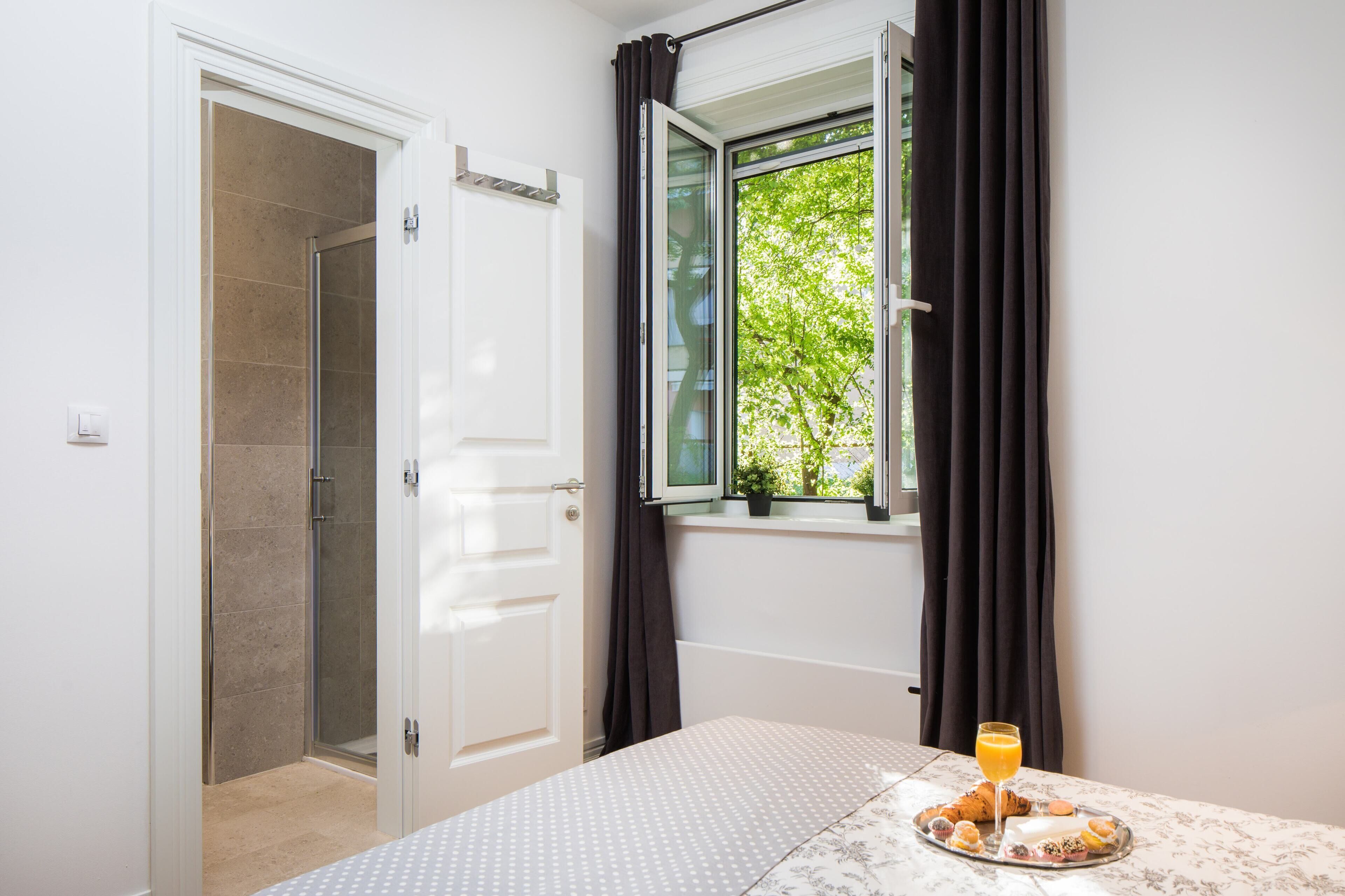 A bedroom with a bed, a window with curtains, a tray with pastries and a glass of orange juice, and a shower area.