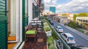 Un balcó amb taula i cadires de fusta, una planta en test i vista d'un carrer amb cotxes i edificis.