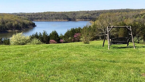 A grassy area with a wooden swing set and a bench overlooking a lake surrounded by trees.
