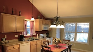A kitchen with wooden cabinets, a dining table with chairs, a chandelier, and a window with a view of trees.