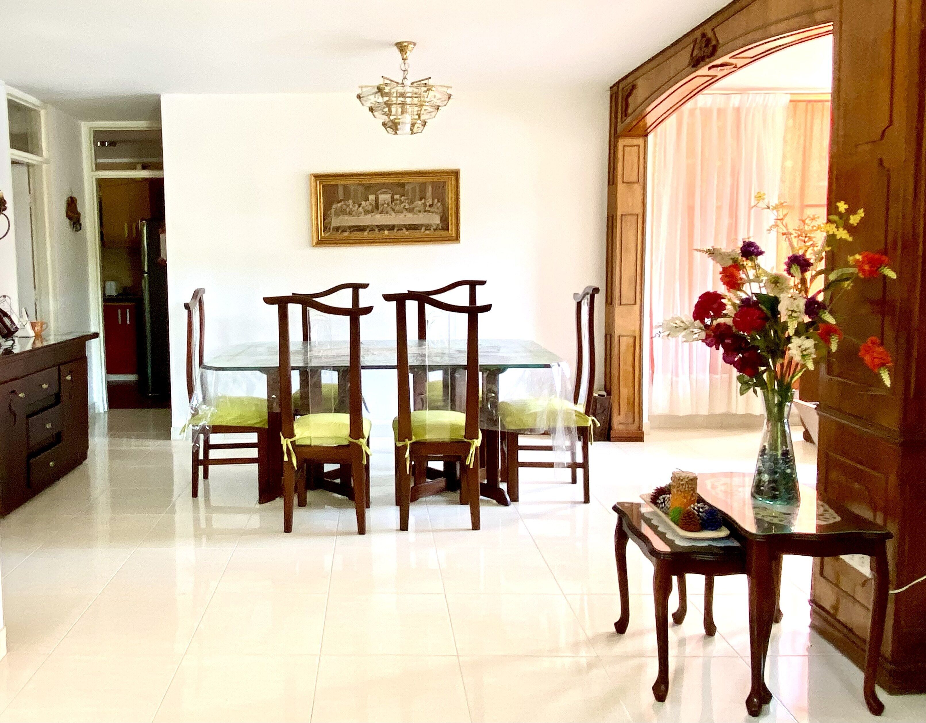 A dining area with a glass table, wooden chairs, a chandelier, and a painting on the wall.