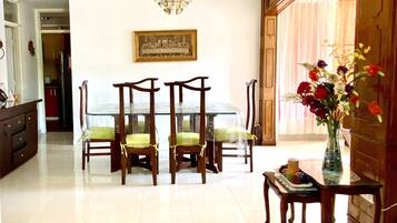 A dining area with a glass table, wooden chairs, a chandelier, and a painting on the wall.
