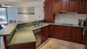 A modern kitchen with wooden cabinets, a granite countertop, a built-in dishwasher, and a view of the outdoors through a large window. - Seas the Day - Lakefront getaway by the beach (Encounter Bay)