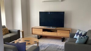 Una sala de estar moderna con un televisor de pantalla plana sobre un mueble de madera, sofás grises, una mesa de centro y un ventilador de techo.