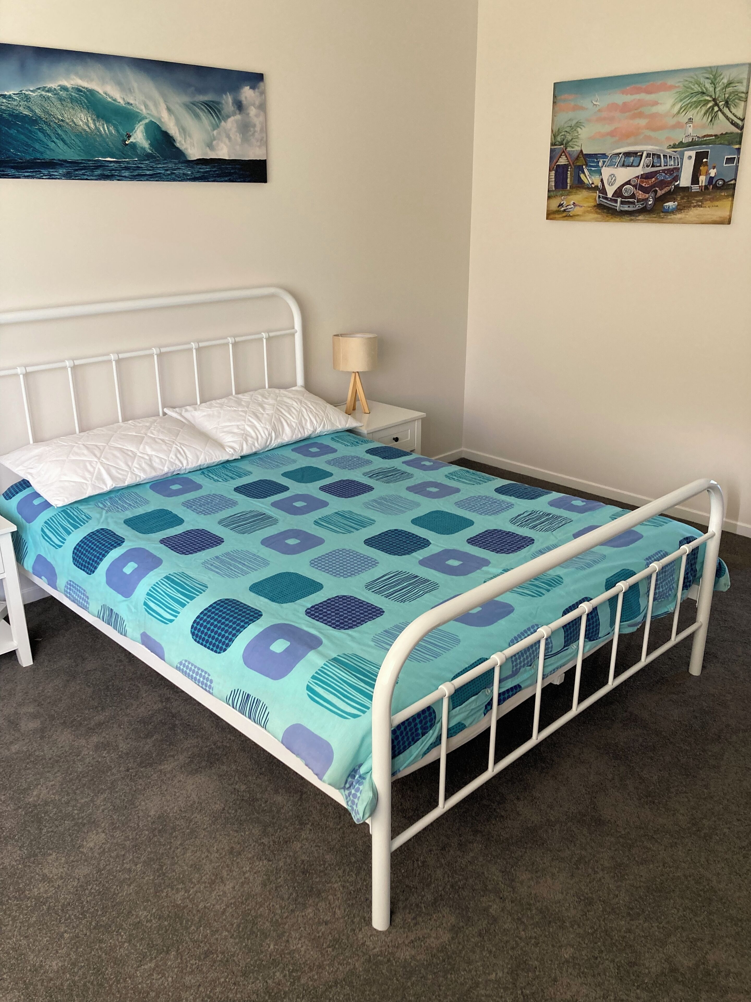 A single-bed room with a metal frame, a patterned bedspread, a bedside table with a lamp, and two wall art pieces.