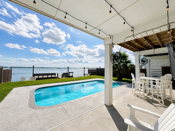A covered outdoor swimming pool with a view of the water, deck chairs, and a fan.