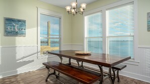 Un comedor con mesa de madera y bancos, ventanas amplias con persianas y una araña.