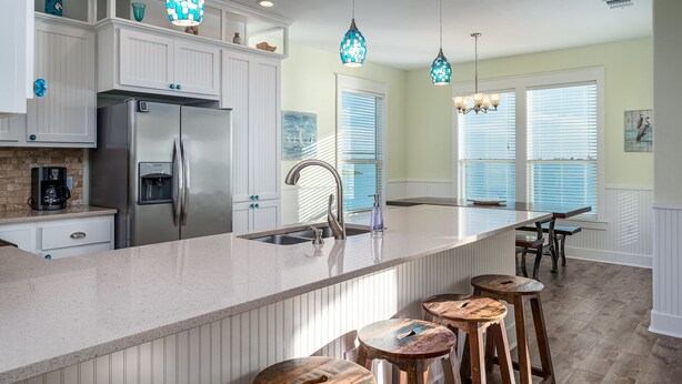 Una cocina con un refrigerador de acero inoxidable, un lavamanos y una isla grande con taburetes. Hay lámparas colgantes sobre la isla.