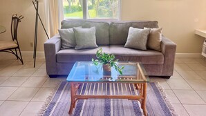 Un salon avec un canapé gris, une table basse en verre, un tapis à motifs, une lampe sur pied et une fenêtre donnant sur des arbres.