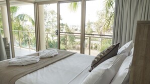 A hotel room with a large bed, a balcony with a railing, and a view of palm trees and buildings. - Le Jarden Vue Du Port - Airlie  Beach (Airlie Beach)
