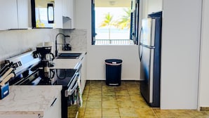 Cocina compacta con estufa, lavamanos y refrigerador, un bote de basura y una ventana con vistas a palmeras.