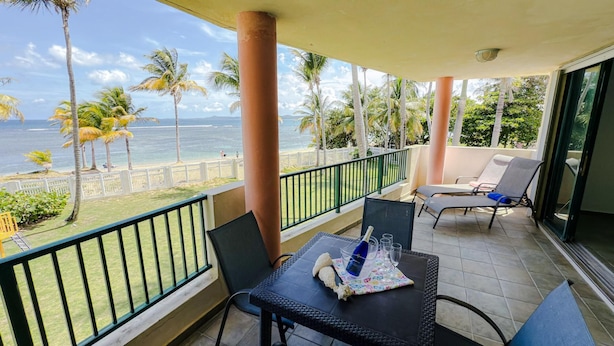 Un balcón con vista al océano, que incluye una mesa con sillas, barandilla, palmeras y la playa.