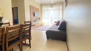 A living room with a dining area, a grey sofa, wooden chairs, a TV on the wall, and a ceiling fan. - Fully furnished apartment with 3 bedrooms and 2 parking spaces. (Prainha)