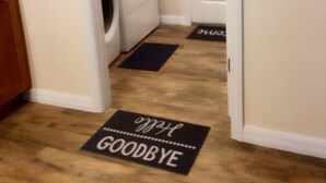 Un lavadero con lavadora y secadora, una puerta con un letrero que dice 'NO OLVIDES' y dos felpudos con la palabra 'ADIÓS'.
