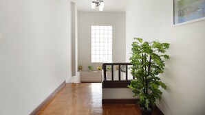 A hallway with wooden flooring, a staircase with a dark railing, and a large potted plant. There is a window with a grid pattern and a ceiling light fixture. - Large 3 Bedroom House Near Old City and Night Bazaar  (Tambon Hai Ya)