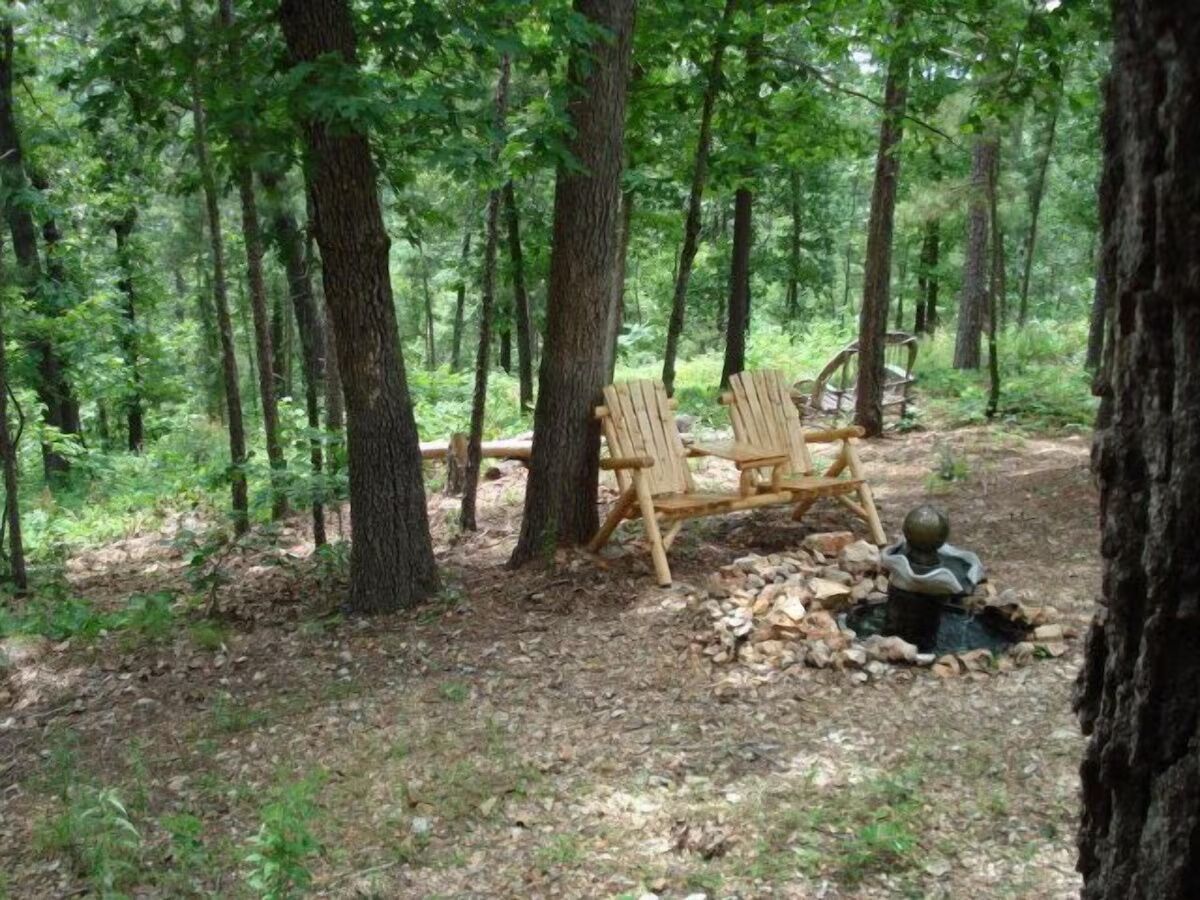 A forested area with two wooden chairs and a small statue near a tree.