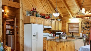 A cozy cabin interior with a wooden ceiling, a central island with a sink, a refrigerator, a microwave, and a dining area with a table and chairs. -  Branson MO Log Cabin Private Porch Quiet Mtn. Forest View Above Lake, Handicap  (Blue Eye)