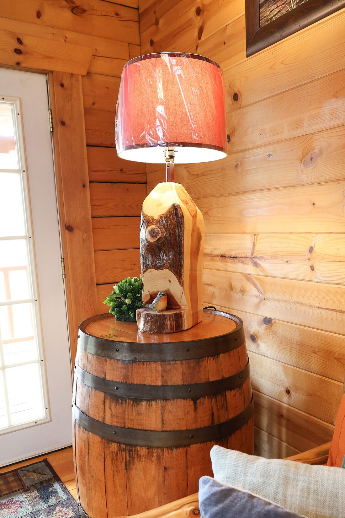A wooden lamp with a red shade on a barrel-shaped base, a small plant, and a cushion on a wooden floor.
