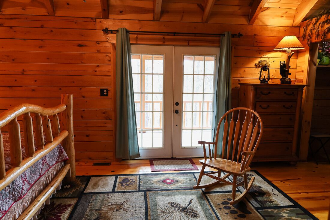 A wooden cabin interior with a rocking chair, a bench, and a lamp on a dresser.