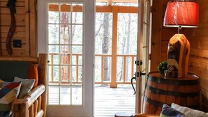 A cozy cabin interior with a wooden floor, a green quilted sofa, a wooden rocking chair, a red lamp, and a large window with a view of trees. -  Branson MO Log Cabin Private Porch Quiet Mtn. Forest View Above Lake, Handicap  (Blue Eye)