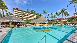 A swimming pool with lounge chairs and thatched-roof structures, surrounded by palm trees and a hotel building. - Marina Sol Los Cabos Haven - Walk to Medano Beach! (Cabo San Lucas)