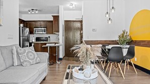 Een moderne woonkamer met een grijze bank, een salontafel met decoratieve objecten, een eethoek met een geel accentmuur, en een keuken met houten kastjes en roestvrijstalen apparatuur.