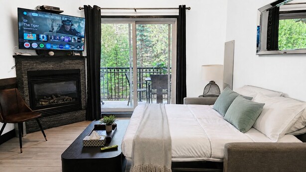 Een slaapkamer met een groot bed, een aan de muur bevestigde televisie, een open haard en een balkon met uitzicht op groen.