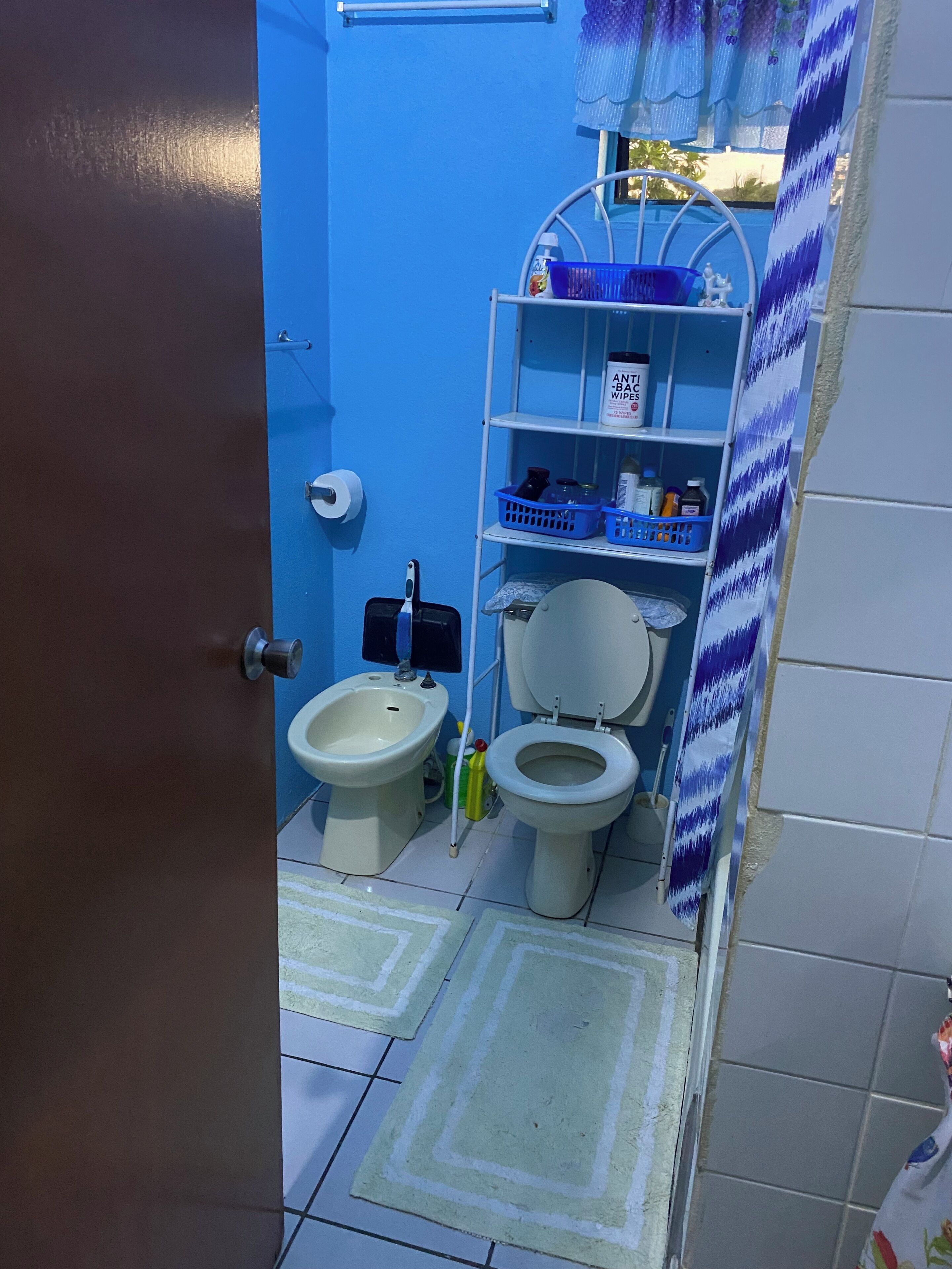 Une salle de bain avec un mur bleu, des toilettes blanches, un bidet et une étagère contenant des articles de toilette.