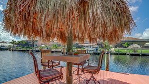 Une terrasse en bois avec une table et des chaises, un parasol en chaume et une vue sur une étendue d’eau, avec des bâtiments en arrière-plan.