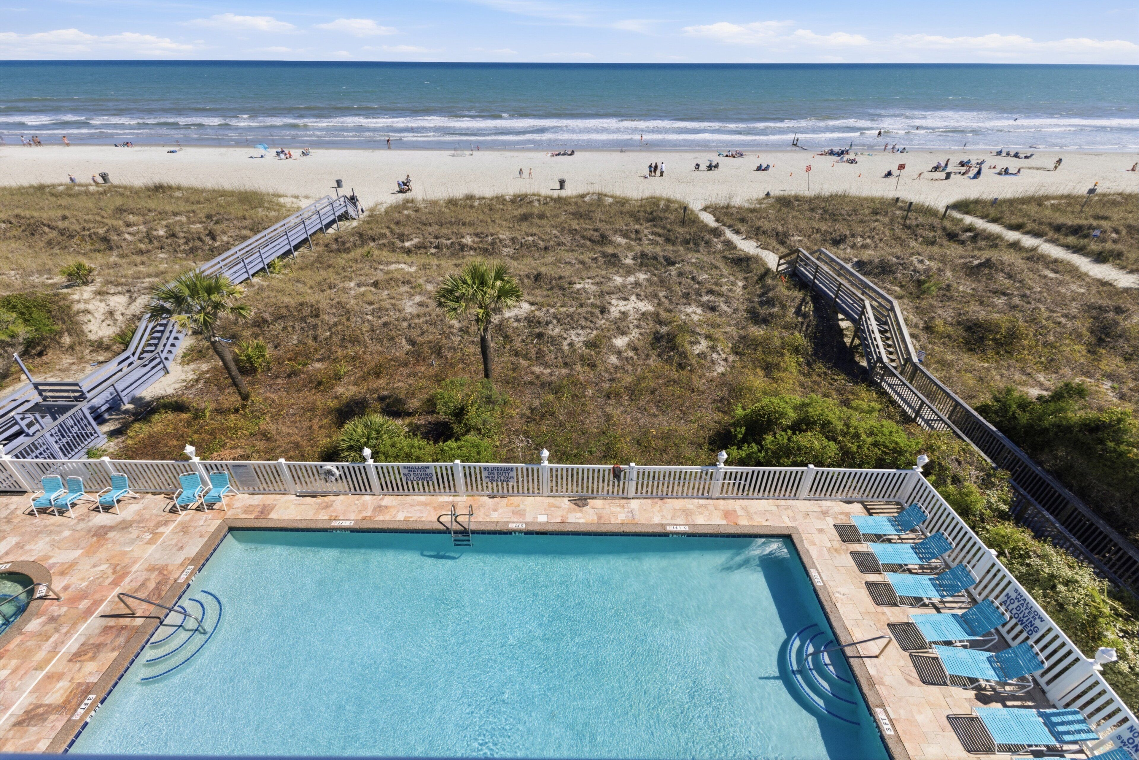 A pool with a view of the beach, a deck with chairs, and a pathway leading to the beach.