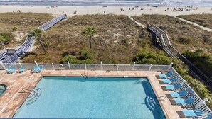 A pool with a view of the beach, a deck with chairs, and a pathway leading to the beach.