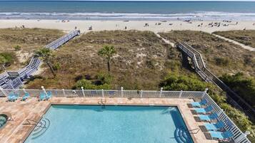 A pool with a view of the beach, a deck with chairs, and a pathway leading to the beach.