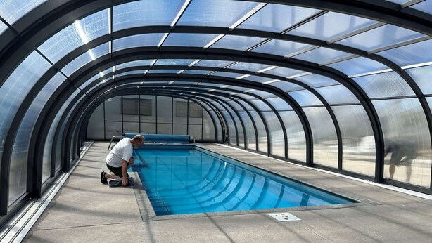 A swimming pool with a curved glass enclosure, a man kneeling by the pool, and a blue sky visible through the structure. - Otorohanga Kiwi Holiday Park (Otorohanga)