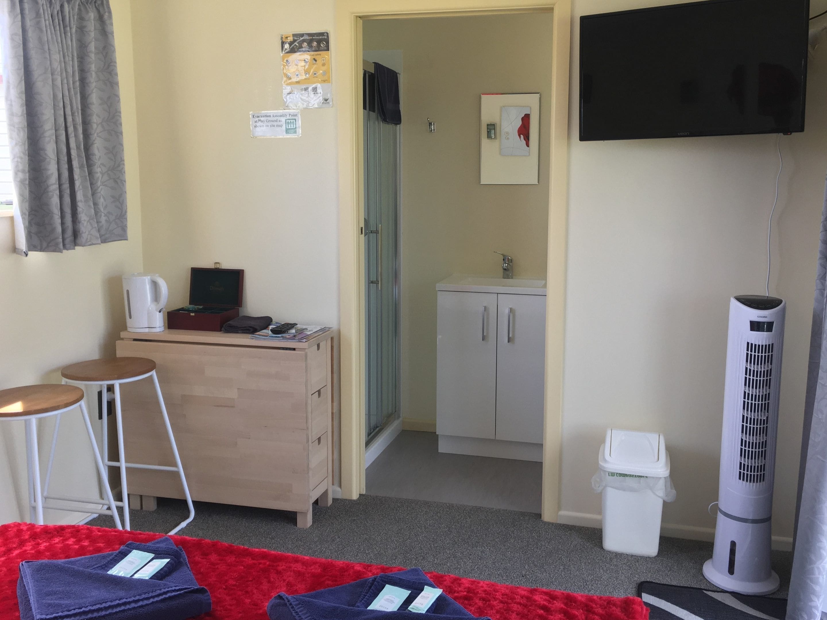 Une pièce compacte comprenant un coin cuisine, un petit tabouret de bar, une télévision, une armoire blanche et un ventilateur sur pied.