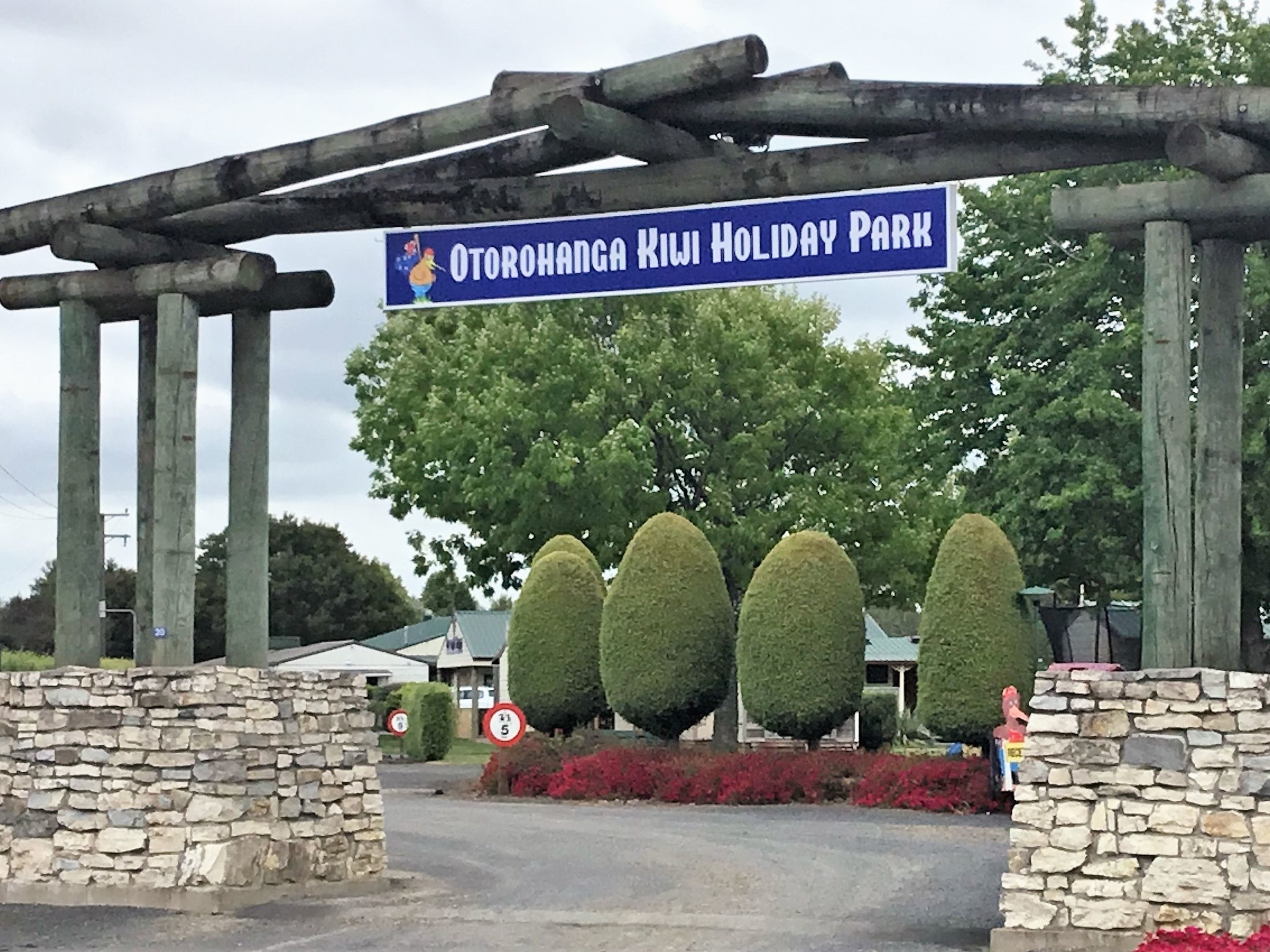 A sign for Otorohanga Kiwi Holiday Park with stone walls and neatly trimmed hedges.