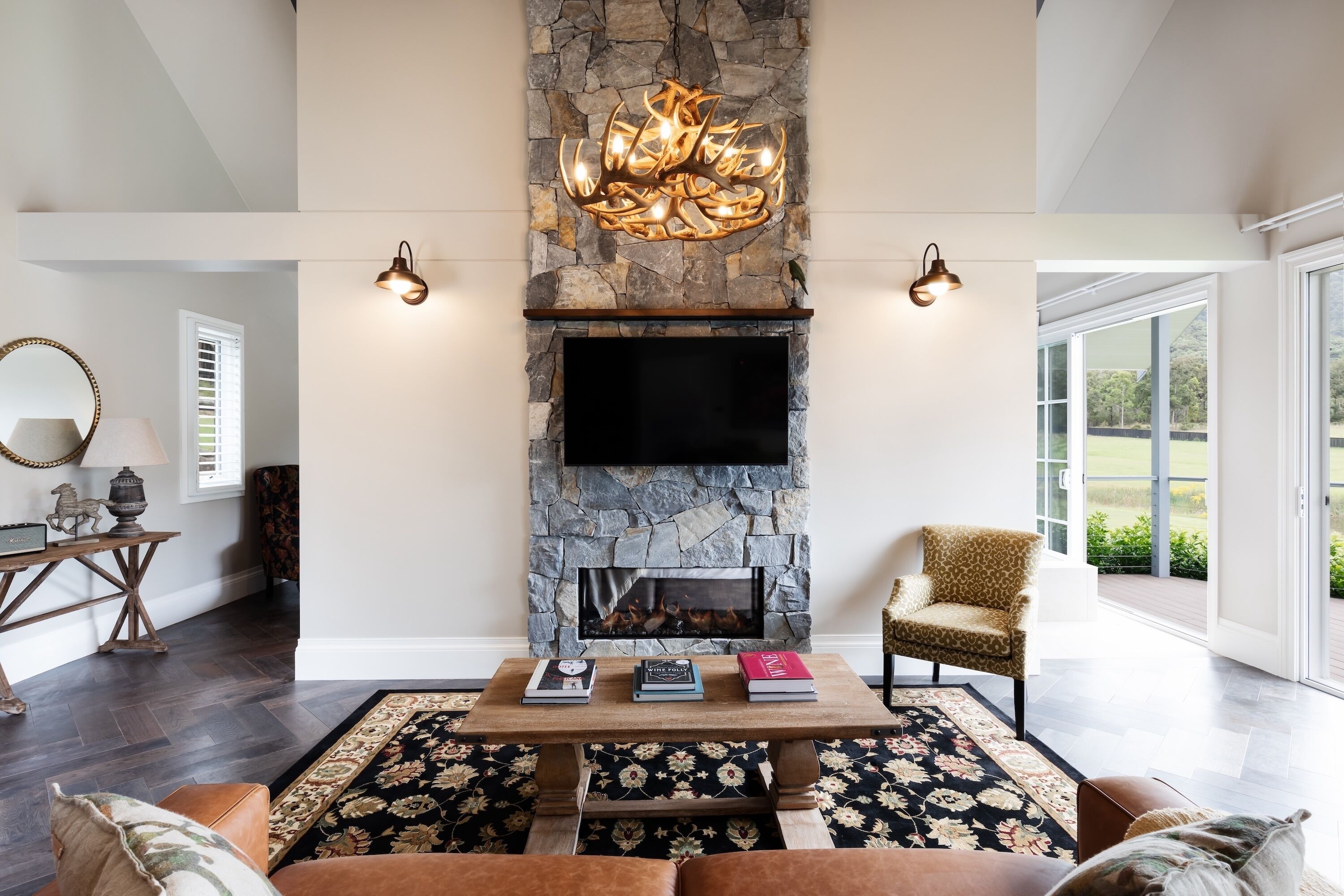 A living room with a stone fireplace, a flat-screen TV, a coffee table with books, a patterned rug, and a leather sofa.