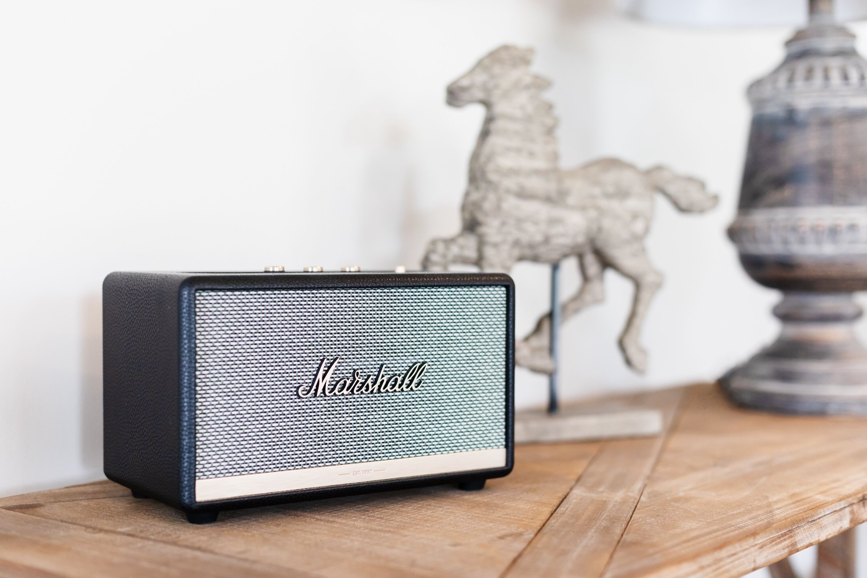A Marshall speaker on a wooden table with a decorative horse and lamp in the background.