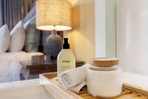 A bathroom with a bottle of Jurlique Restoring Shower Gel, a rolled towel, and a jar of cream on a wooden tray.
