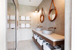 A modern bathroom with a glass shower enclosure, two sinks with round mirrors, and built-in shelves with towels.