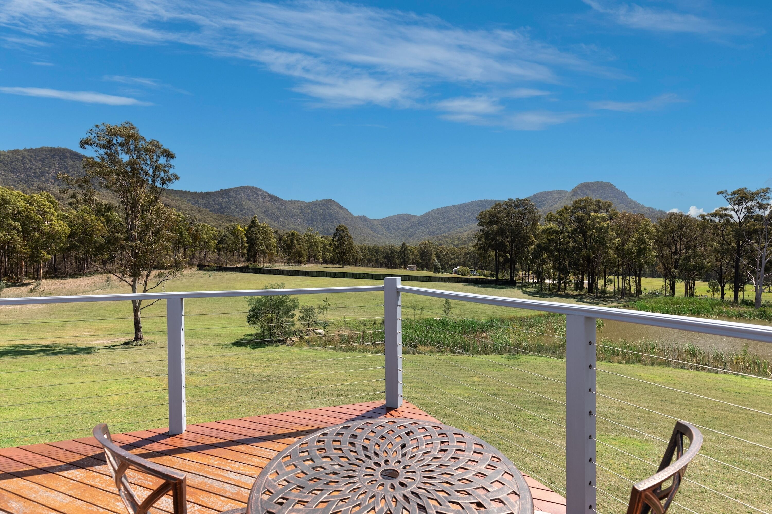 A deck with a metal table and chairs, overlooking a grassy field and hills.