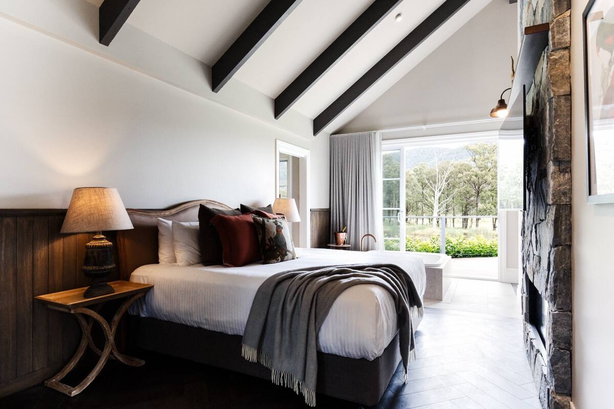 A bedroom with a large bed, bedside lamps, a view of the outdoors, and a stone fireplace.