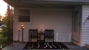 Un portico con due sedie di legno e un tavolino, un tappeto decorativo e due candelabri con candele.