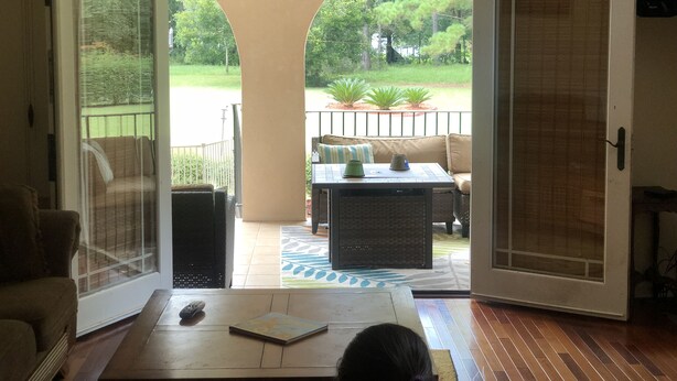 Una puerta corredera abierta que da a un patio con un ventilador de techo, una zona de estar con cojines y una pequeña mesa con un control remoto y un libro encima.