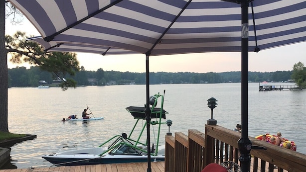 Une terrasse avec des chaises longues rouges et un parasol à rayures, donnant sur un lac paisible où l’on peut voir des bateaux et des personnes dans l’eau.