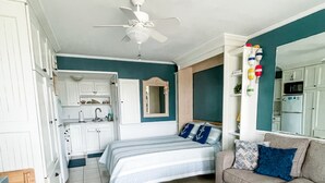 A compact living space with a kitchenette, a bed with striped bedding, a sofa with decorative pillows, and a ceiling fan.