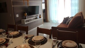 A dining area with a table set for four, a television on a wooden stand, and a living area with a sofa and curtains.