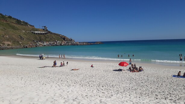 A beach with people sunbathing and swimming, a red umbrella, and a building on a hill in the background.