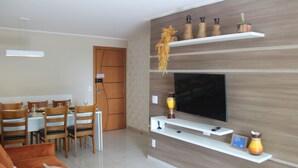 A modern living room with a flat-screen TV, wooden shelves with decorative items, a dining area with a table and chairs, and a sofa.