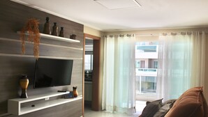 A modern living room with a flat-screen TV, built-in shelves, a brown sofa, and a large window with sheer curtains.